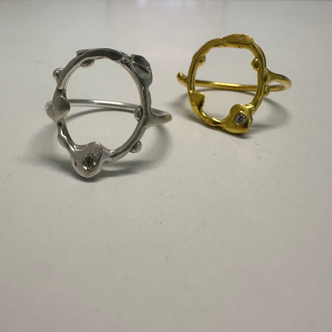 Two abstract open ring rings with organic wavy edges, one in silver and one in yellow gold, each featuring a small clear gemstone, displayed on a white surface.