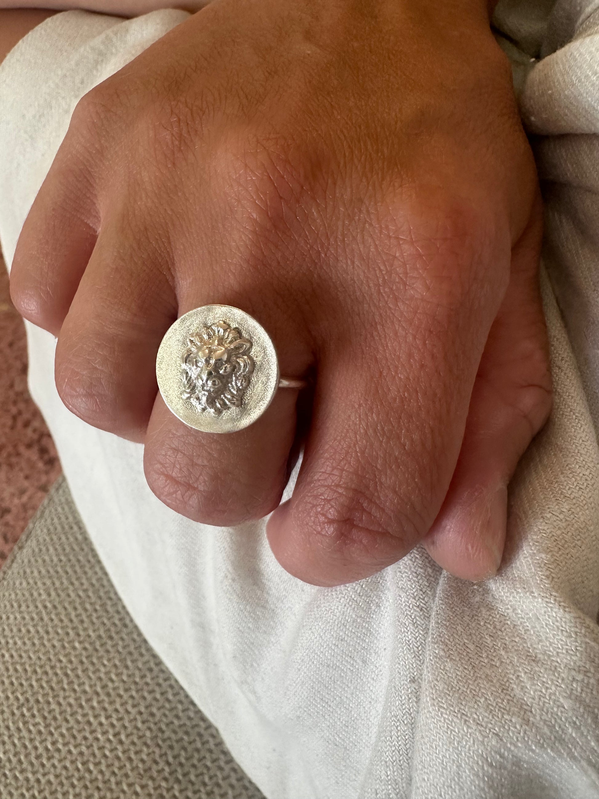 A close-up of a hand wearing a silver signet ring with an embossed lion head design, resting on light-colored fabric.