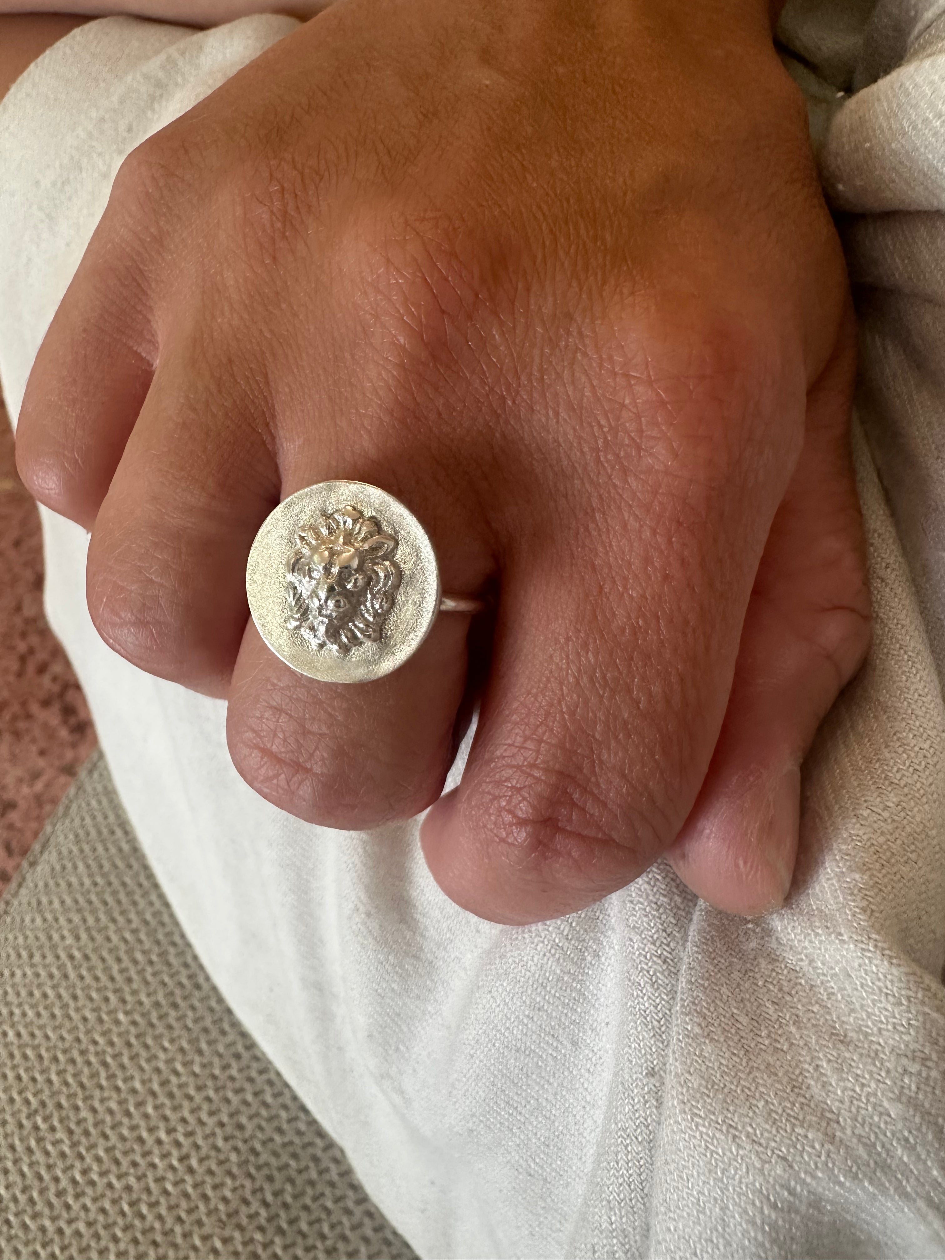 A close-up of a hand wearing a silver signet ring with an embossed lion head design, resting on light-colored fabric.