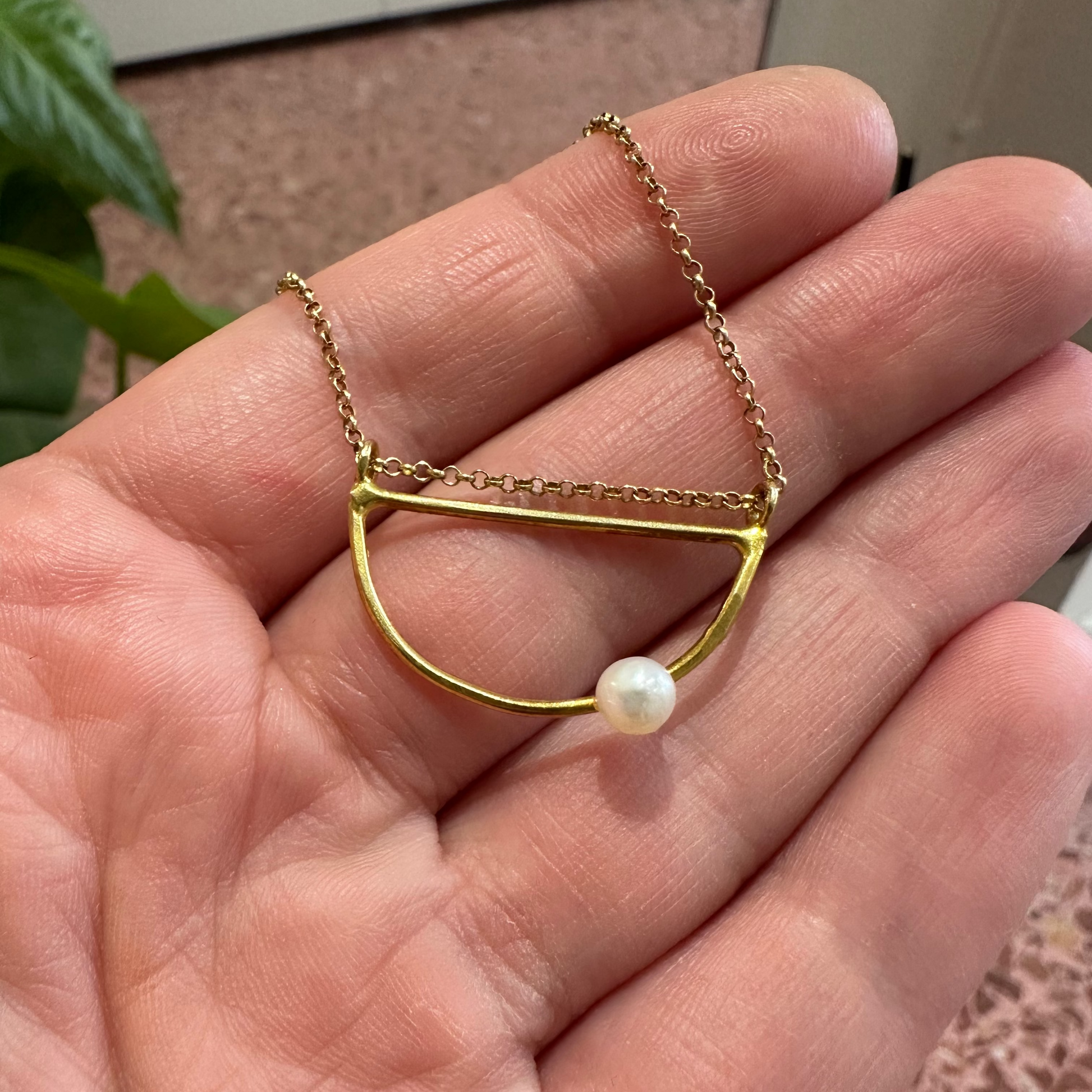 A gold necklace with a minimalist semi-circular bar and a single white pearl pendant, held in a person's hand.