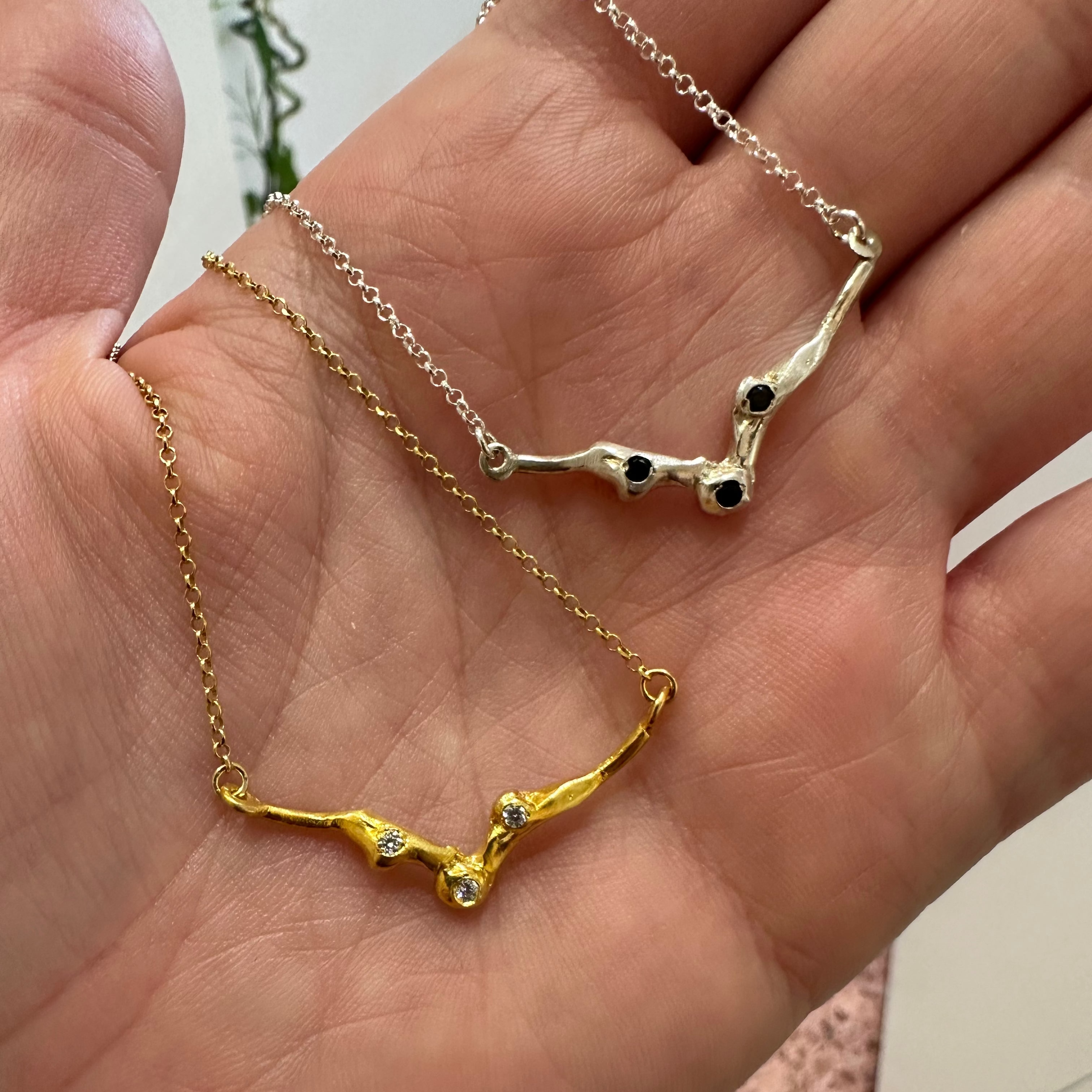Two delicate tree branch-style necklaces, one gold and one silver, each with small black and clear stones, held in a person's hand.