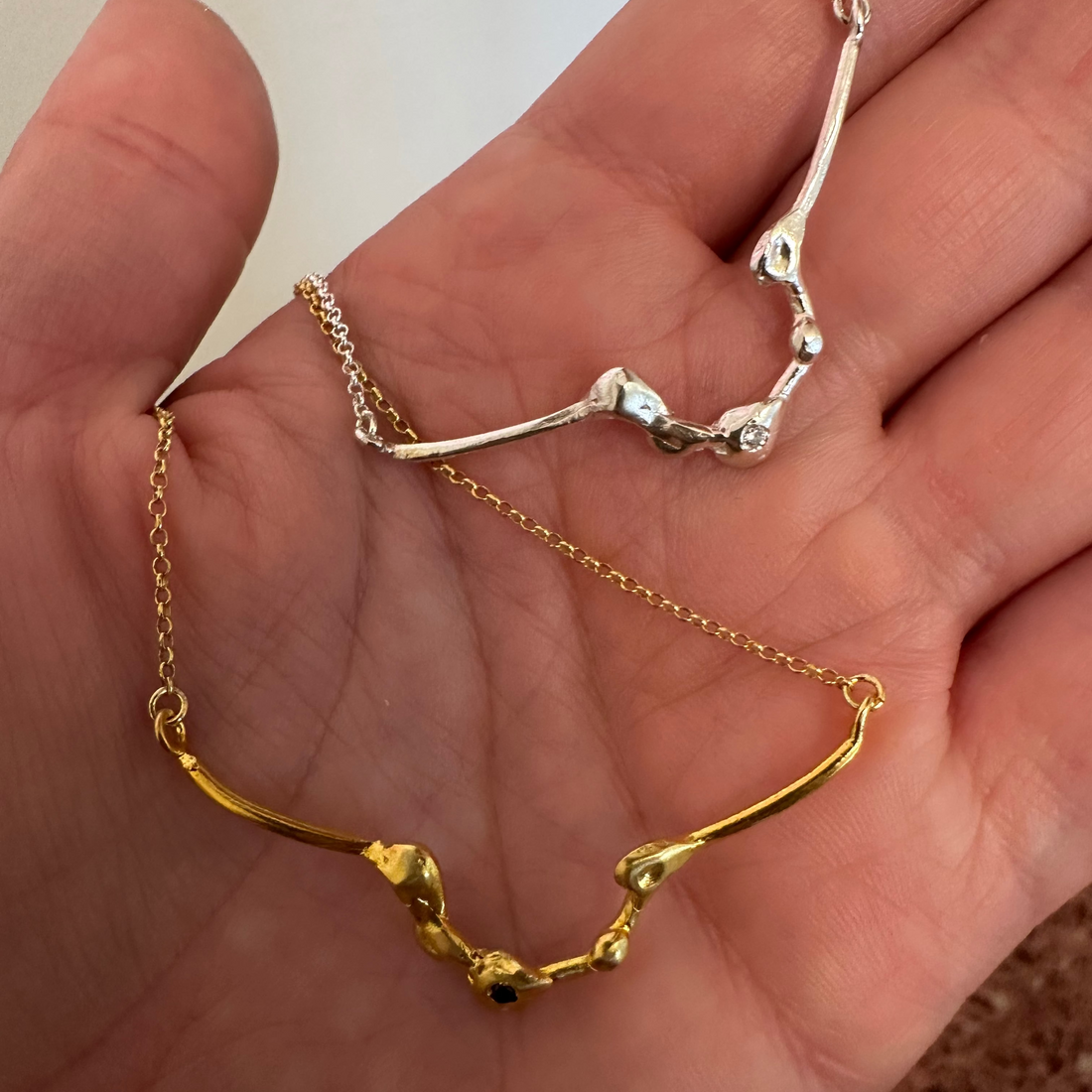 A gold necklace featuring a textured, organic-shaped pendant with a small dark blue gemstone accent at the center, displayed on a clean white background.
