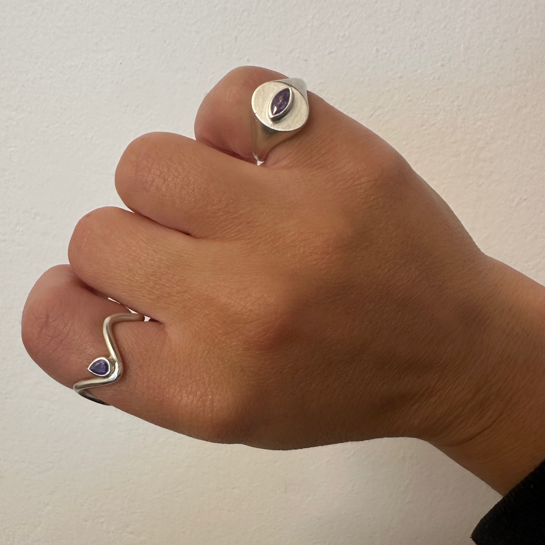 A close-up of a hand wearing two silver rings: one with a flat oval setting and a small dark oval stone, and another with a wavy open band design and a small dark heart-shaped stone.