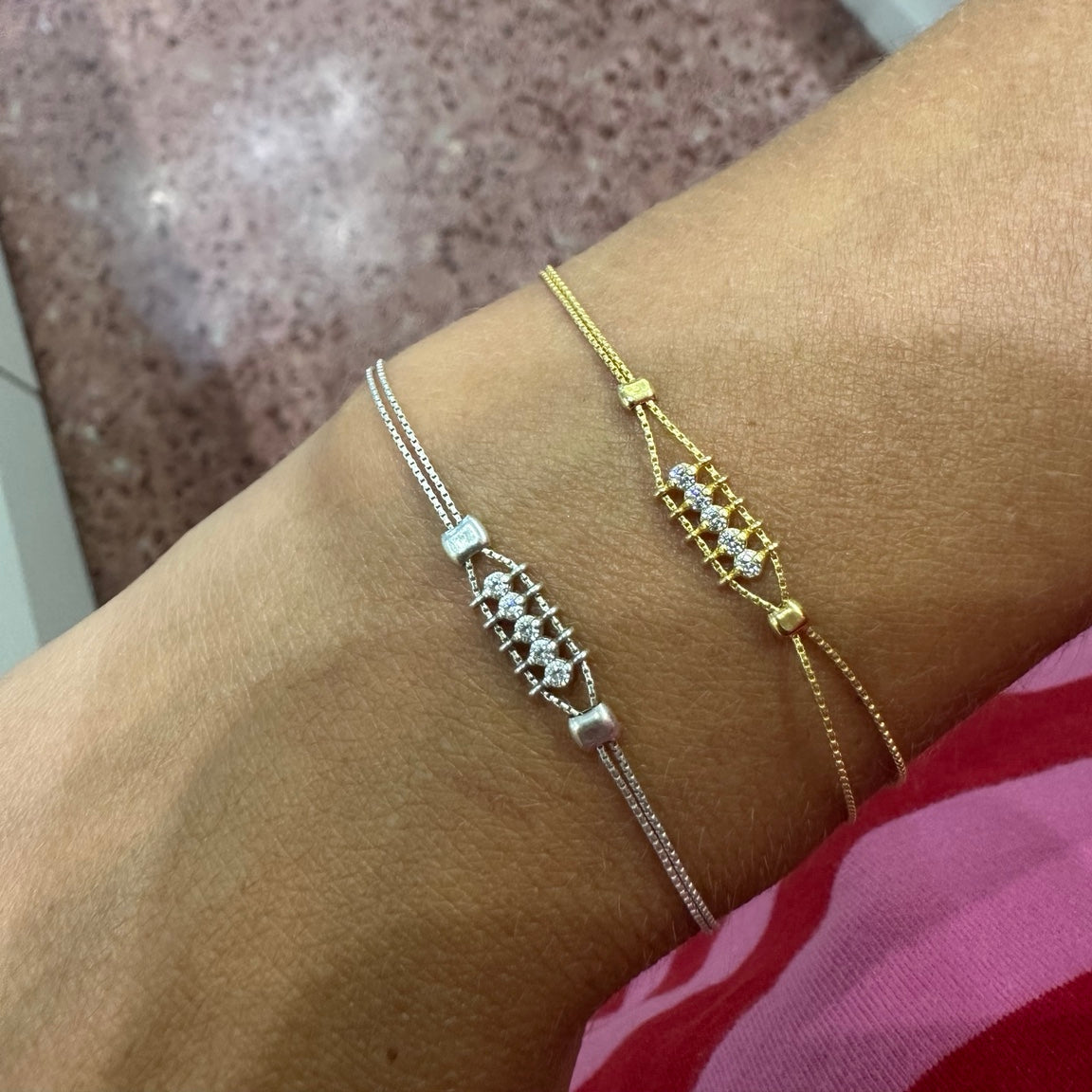 Two delicate bracelet-style wristlets with small clear stones are shown worn on a person's wrist. One is silver-toned and one is gold-toned, photographed indoors against a pink speckled floor and white furniture.