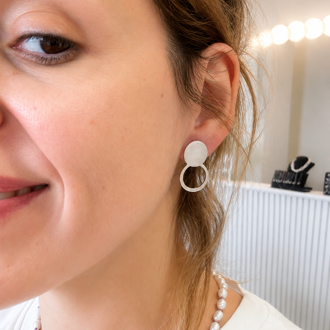 A close-up of a woman wearing a minimalist double-ring drop earring in white, with her hair pulled back and a pearl necklace visible.