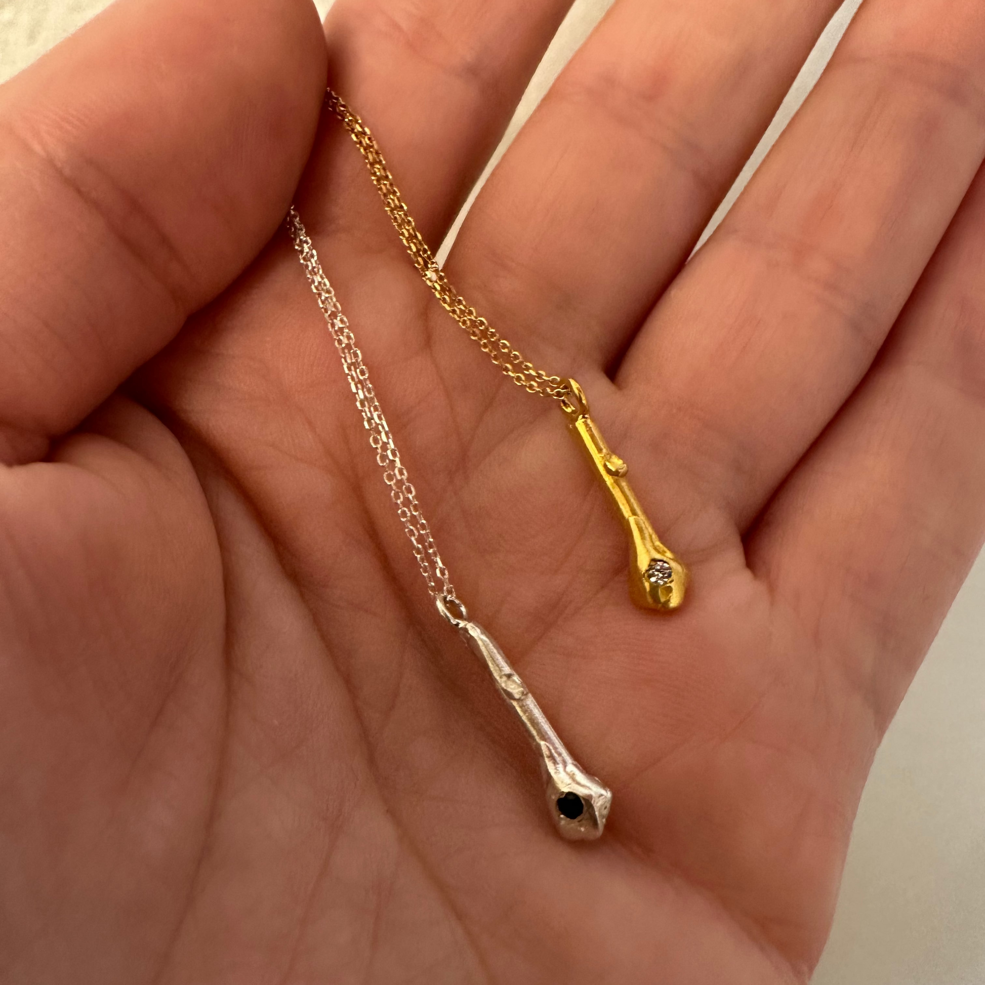 A close-up of a person's hand holding two twig-style pendant necklaces, one gold and one silver, each with a small dark gemstone.