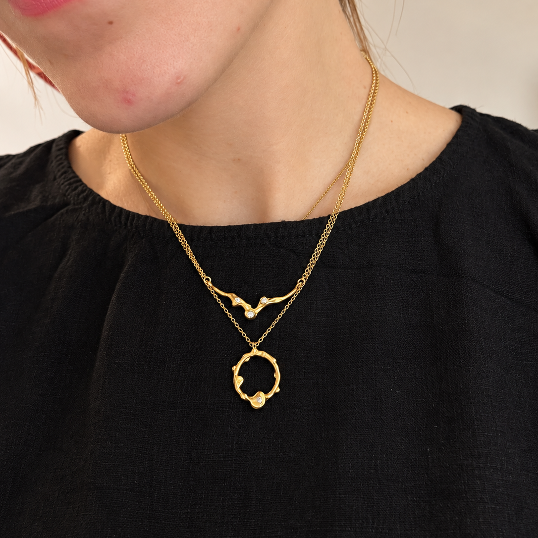 A close-up of a person wearing a delicate gold necklace with a double-layered design, featuring a small wave-like pendant and a textured circular ring with a bead, over a black top.