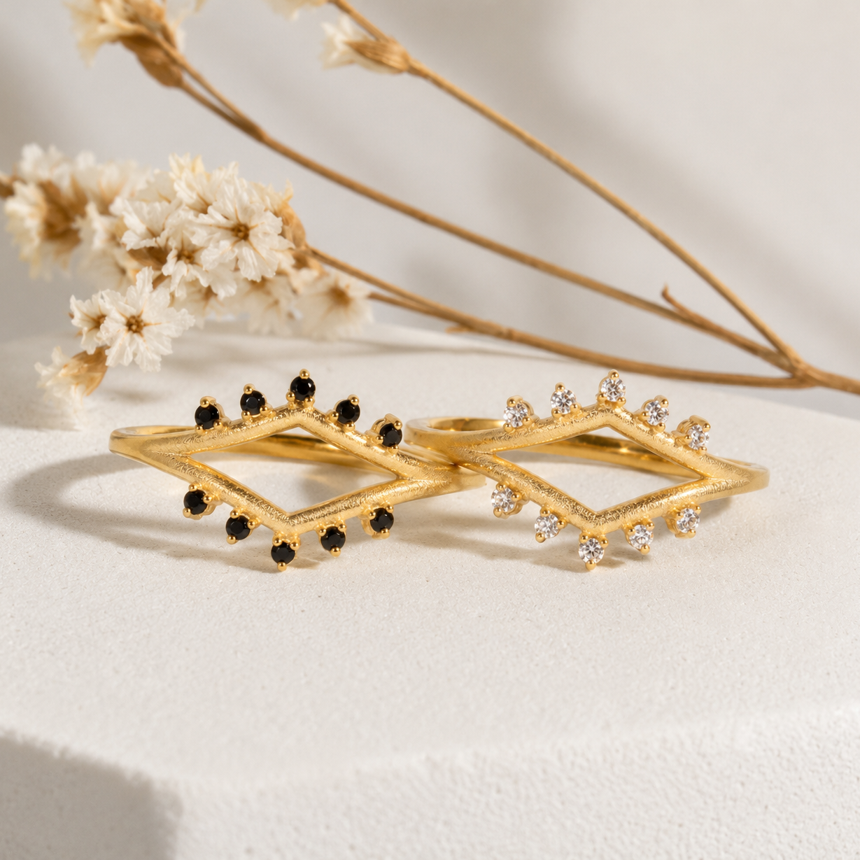 Gold and black diamond-shaped earrings on a neutral background with dried flowers.