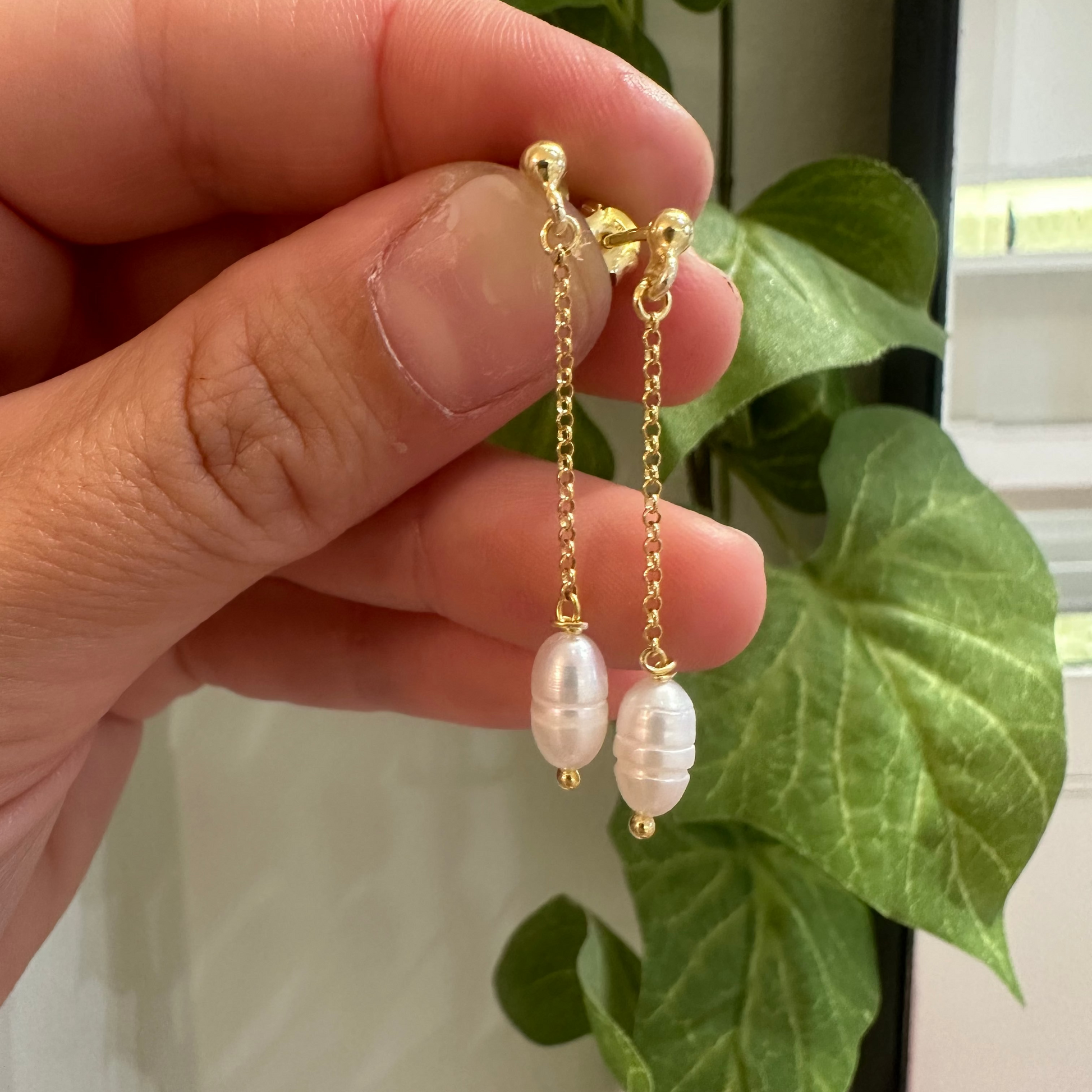 Gold chain earrings with pearl drops held by a hand in front of green leaves.