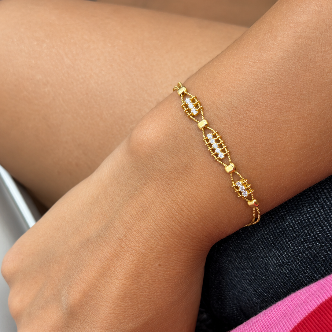 A close-up of a person's wrist wearing a delicate gold bracelet with repeating oval links set with small clear stones, resting against dark denim and a pink garment.